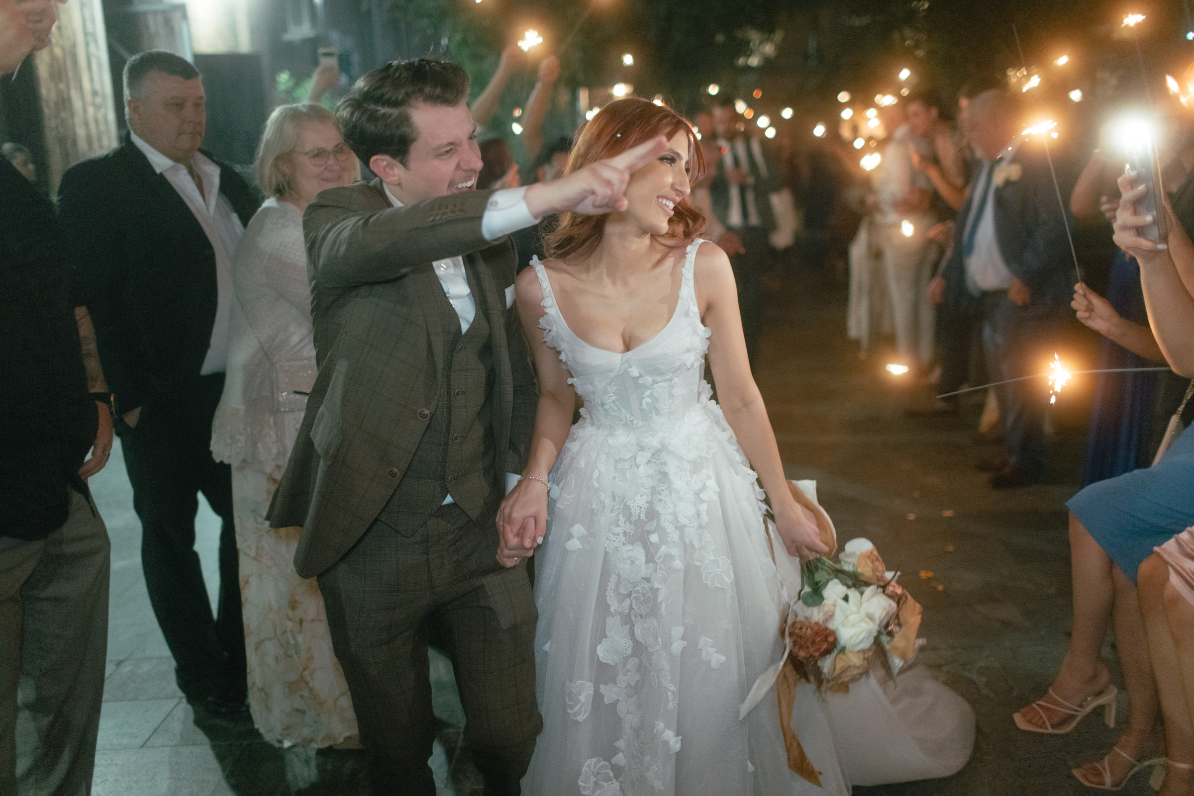 A couple leaves their wedding with all of their guests holding up long sparklers. The wedding photo shows the groom pointing saying goodbye to his guests.